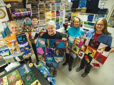 This photo shows five ladies holding lap sized fidget quilts made with various designs. Behind them is the sewing workshop where the quilts are made.